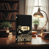 A black personal finance budget binder with a gold lock is displayed on a wooden desk next to a lamp and books.