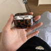 A hand holds a glossy tortoiseshell protective case for AirPods against a striped background.