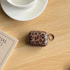 A small, rounded earphone case cover with a leopard print design rests on a wooden surface next to a white cup and saucer.