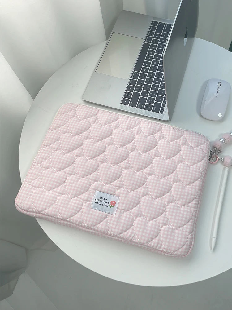 A pink quilted laptop sleeve with a checkered pattern resting on a white table next to a silver laptop.