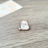 Un pequeño broche de esmalte con una gata blanca sonriente sobre una superficie de madera.