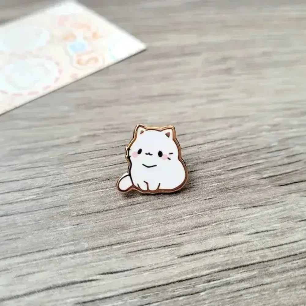Un pequeño broche de esmalte con una gata blanca sonriente sobre una superficie de madera.