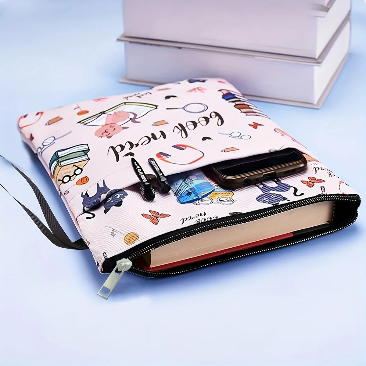 A pink fabric book sleeve with a zipper and various stationery items is shown resting on a light blue surface.