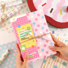 Manos sostienen un papel de scrapbook rosa cuadriculado con puntos y texto colorido, junto a una base roja.