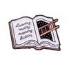 An enamel lapel pin shaped like an open book with the text Leaving reality entering fiction.