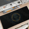 A black gaming mouse pad with gold Lord of the Rings themed design is displayed on a wooden desk next to a keyboard and mouse