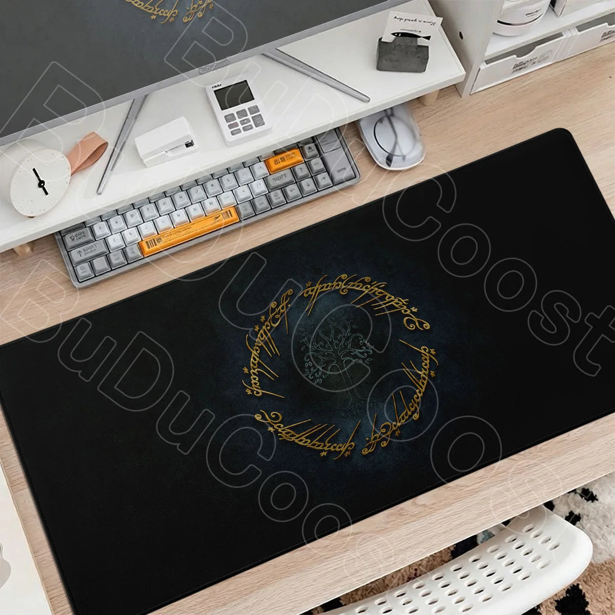 A black gaming mouse pad with gold Lord of the Rings themed design is displayed on a wooden desk next to a keyboard and mouse