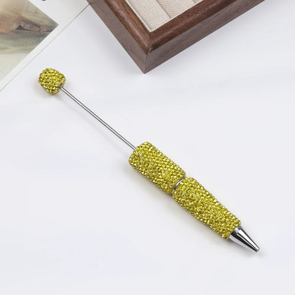 A gold-colored pen accessory featuring a rhinestone-encrusted tip and body, displayed on a white surface.