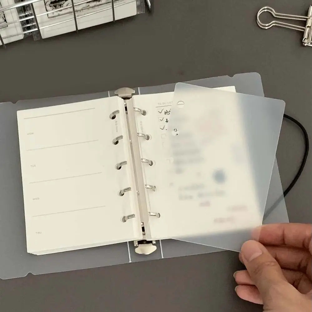 A hand holds open a transparent divider page for loose-leaf planners, showing organized sections and checkmarks.
