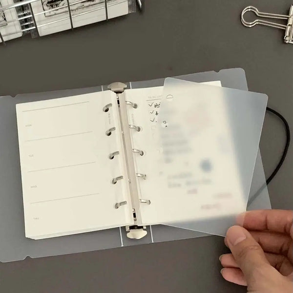 A hand holds open a transparent divider page for loose-leaf planners, showing organized sections and checkmarks.