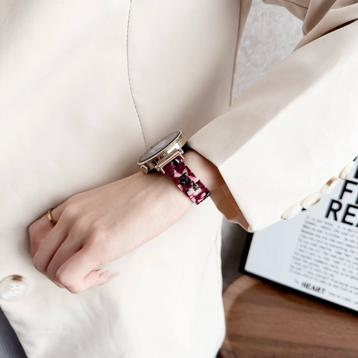 A close-up of a wrist wearing a gold watch with a red and black resin beaded watch band, shown against a light blazer.