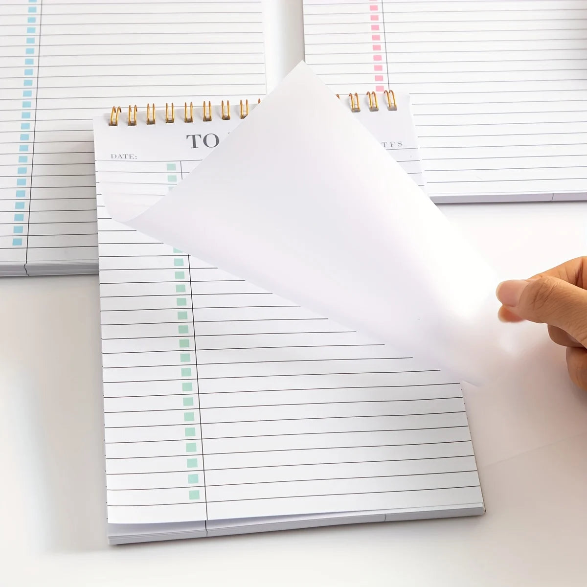 Una mano sostiene una hoja de papel blanco, que oculta parcialmente un bloc de notas diario sin fecha con líneas rayadas.