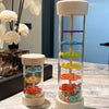 A clear glass tube filled with colorful plastic beads, mounted on a wooden base and held by a wooden top, used as a musical i
