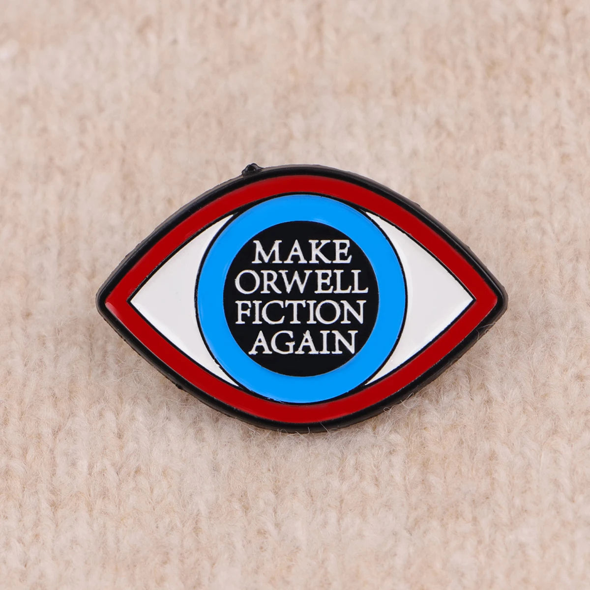 An enamel lapel pin shaped like an eye with the text MAKE ORWELL FICTION AGAIN inside.