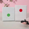 Two square labels, one silver with a green dot and one white with a red dot, are displayed on a light surface.