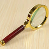 A gold-framed magnifying glass with a reddish wooden handle is shown on a light wooden surface.