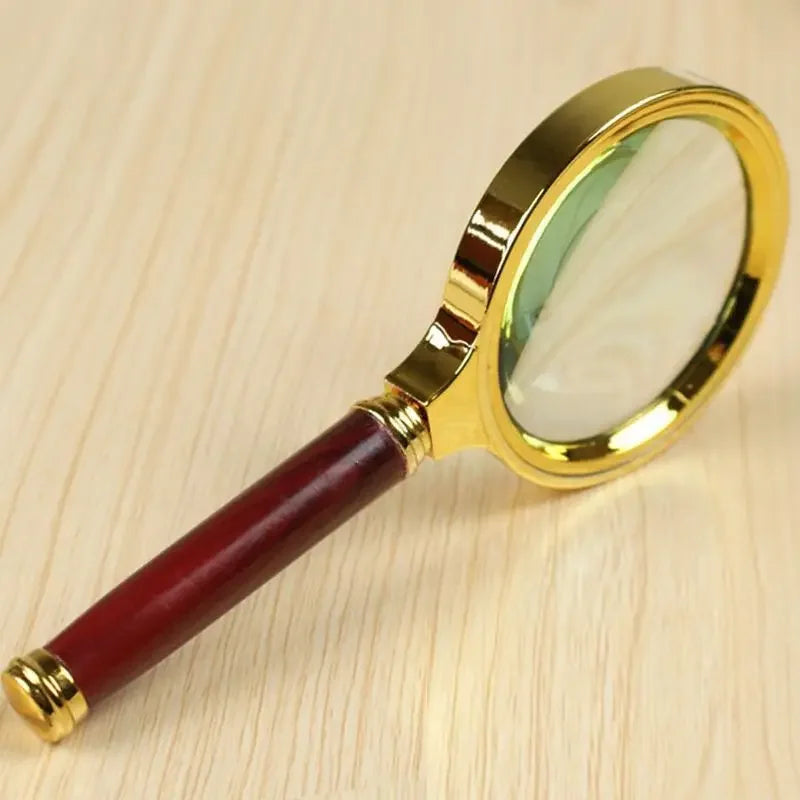 A gold-framed magnifying glass with a reddish wooden handle is shown on a light wooden surface.
