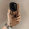 A hand holds a phone case featuring a brown spider web design and a black back, with a silver watch visible.
