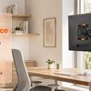 A black fully adjustable monitor arm is mounted on a wooden desk next to a window.