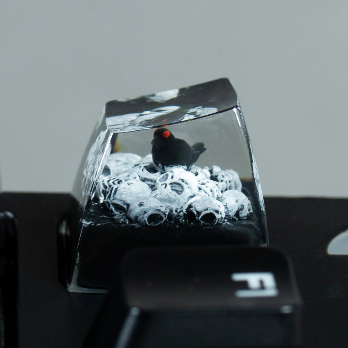 A black resin keycap featuring a gothic raven and skull design, resting on a dark surface.