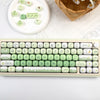 A close-up of a light green mechanical keyboard with custom keycaps and a small woven basket in the background.