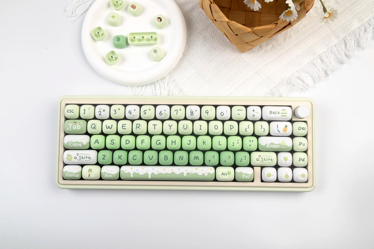 A close-up of a light green mechanical keyboard with custom keycaps and a small woven basket in the background.