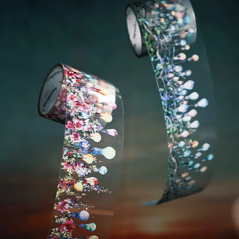 A roll of decorative clear PET washi tape featuring floral and botanical elements, shown in a close-up against a soft, blurre