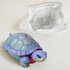 A purple and green turtle toy rests next to its clear plastic shell mold.
