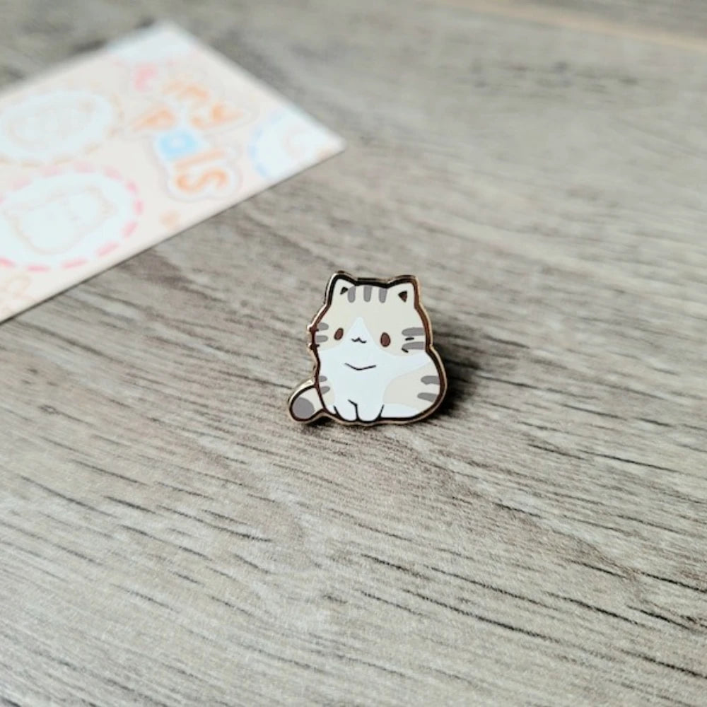 Un pequeño broche de esmalte con forma de un gato sonriente, pelaje blanco y detalles naranjas, sobre una superficie de mader