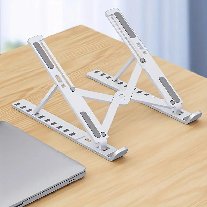 A white adjustable foldable laptop cooling stand is shown resting on a wooden table next to a silver laptop.
