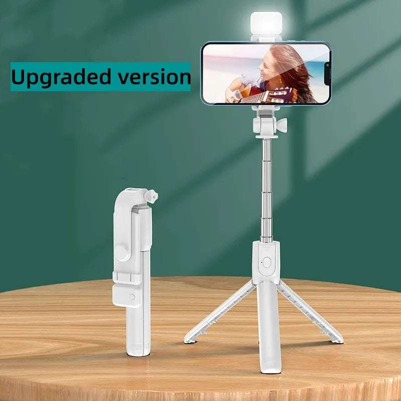 A white selfie stick tripod with a fill light and wireless remote is displayed on a wooden table.