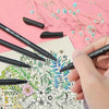 A hand holds a black fineliner pen writing on paper, with other pens and delicate floral branches in the background.