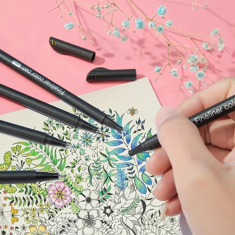 A hand holds a black fineliner pen writing on paper, with other pens and delicate floral branches in the background.