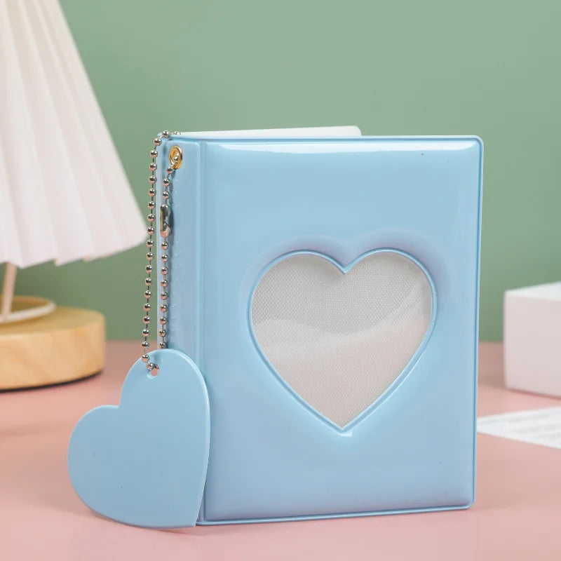 A light blue hollow photo card binder with a heart-shaped cutout and a dangling charm resting on a pink surface.