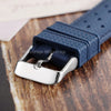 A close-up of a dark blue perforated silicone watch strap with a silver quick release buckle resting on a wooden surface.