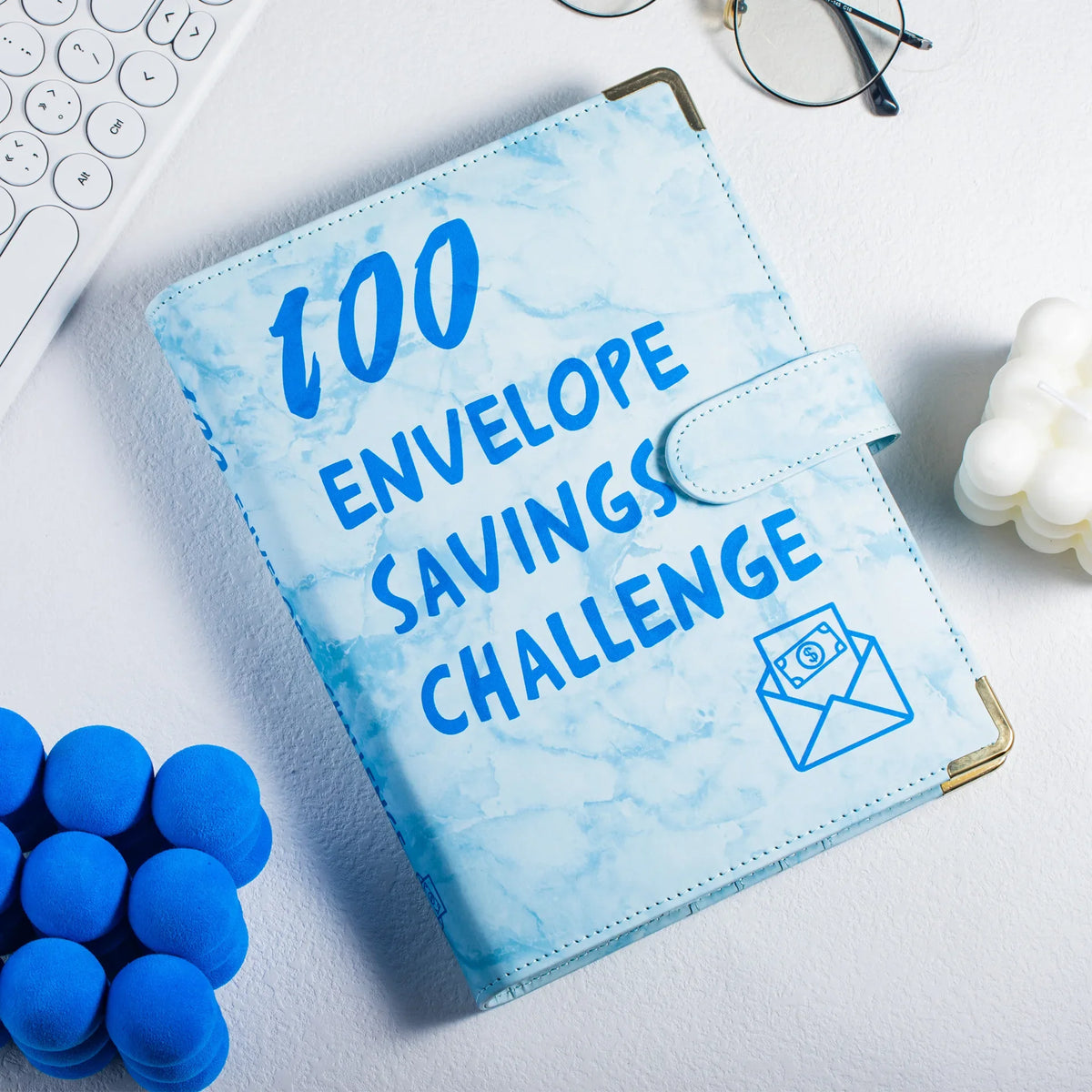 A blue marble budget binder titled 100 Envelope Savings Challenge resting on a white surface.