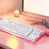 A pink acrylic keyboard riser stand with an ergonomic wrist rest is placed on a light pink desk next to a monitor and a candl