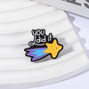 A black enamel pin shaped like a star with a yellow face and blue wings, featuring the text you did it.