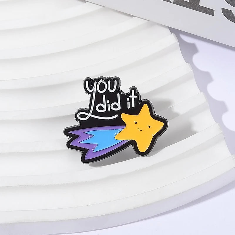 A black enamel pin shaped like a star with a yellow face and blue wings, featuring the text you did it.