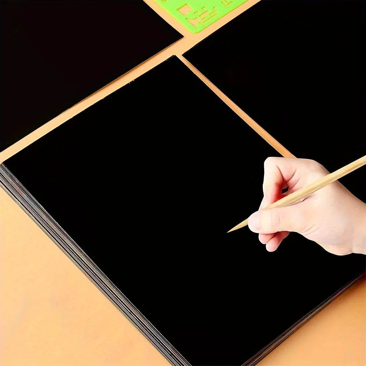 A hand is using a stylus to draw on a black scratch-off art paper sheet.