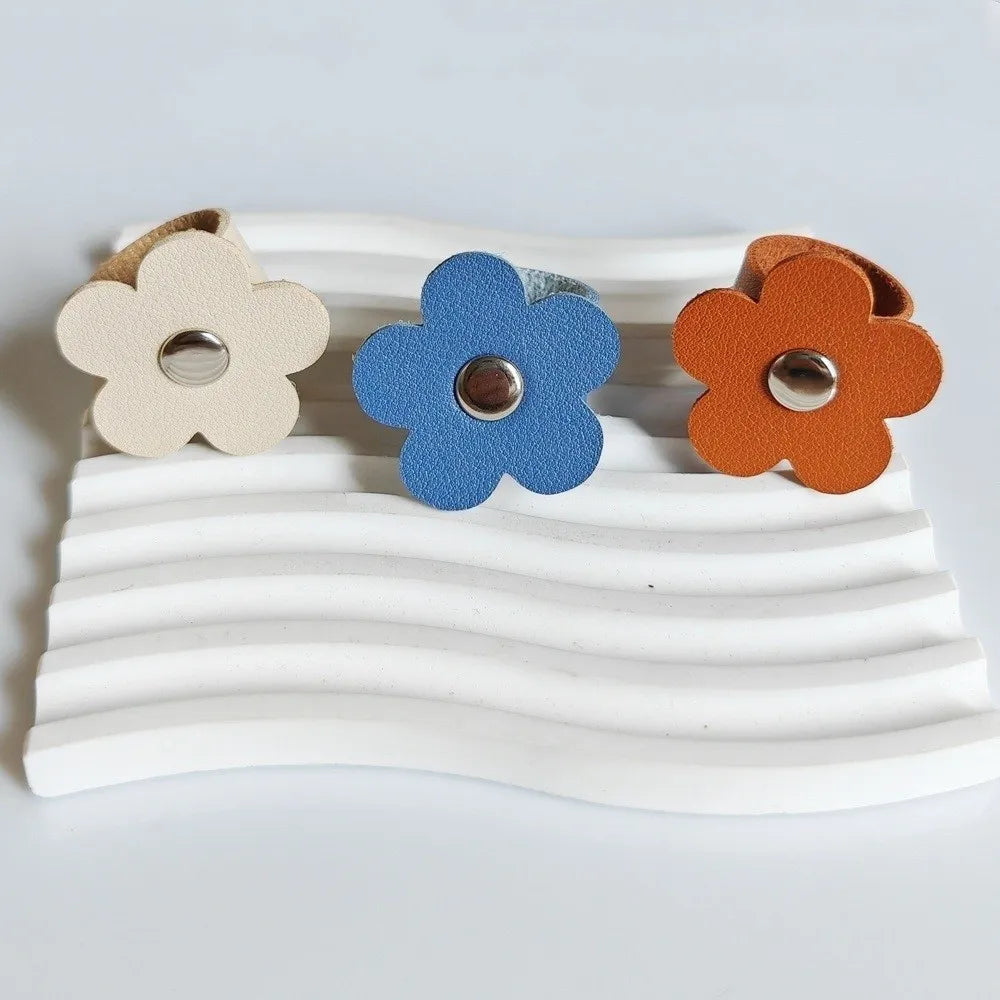 Three leather flower cable organizers in beige, blue, and orange resting on a white wavy surface.