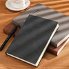 A dark gray notebook is placed on a wooden surface next to a pen and a small white cup.
