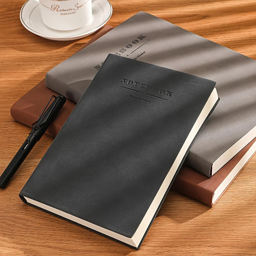 A dark gray notebook is placed on a wooden surface next to a pen and a small white cup.