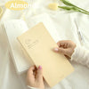 A hand holds a beige slim weekly planner journal next to some almonds and green leaves on a white surface.