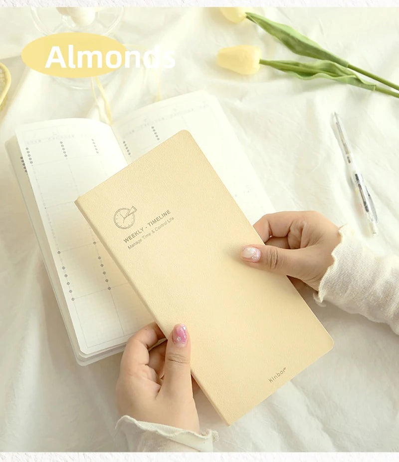 A hand holds a beige slim weekly planner journal next to some almonds and green leaves on a white surface.