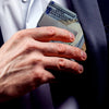 A hand is holding a stack of US dollar bills against a dark suit jacket and a light-colored shirt.