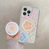 A hand holds a clear floral magnetic phone case for an iPhone featuring pastel colored clouds and a smiling face design.