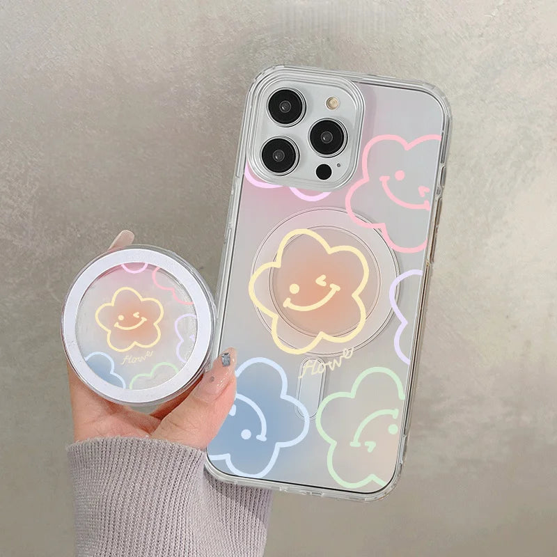 A hand holds a clear floral magnetic phone case for an iPhone featuring pastel colored clouds and a smiling face design.