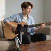 A young man sits on a couch playing an acoustic guitar with an adjustable aluminum tablet stand.