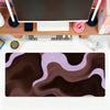 A dark brown and purple abstract wave mouse pad placed on a wooden desk with a laptop and peripherals.